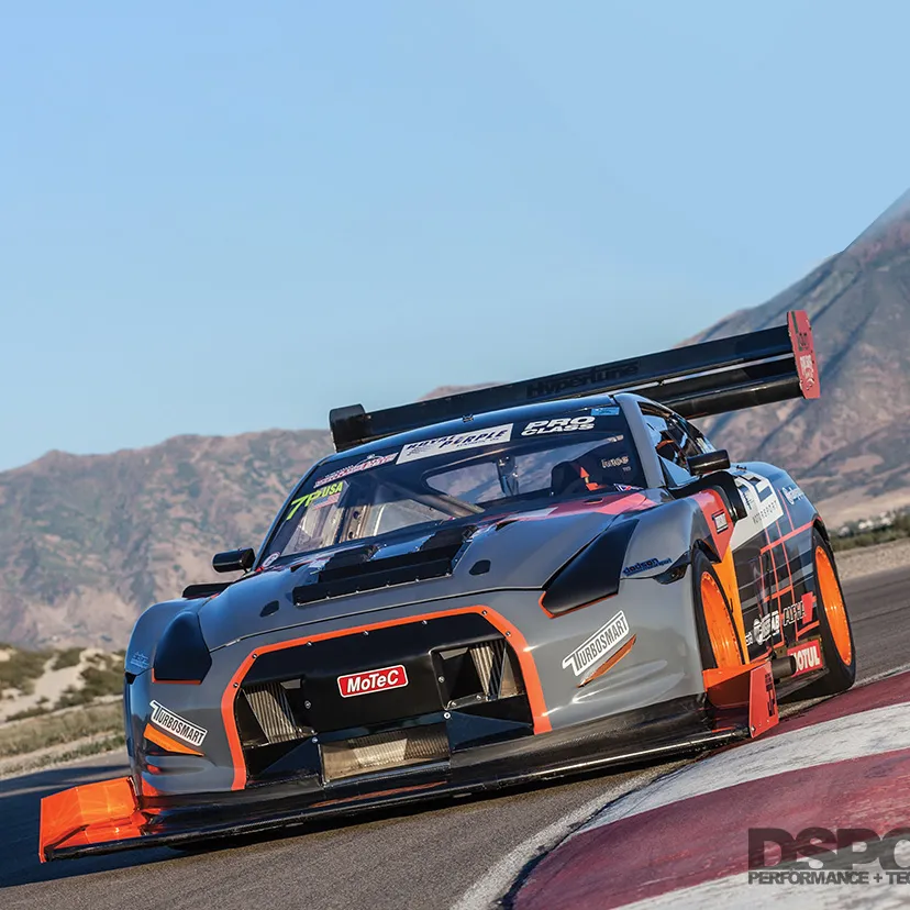 Gray and orange race car with large rear wing navigating a racetrack curve with mountains in the background.