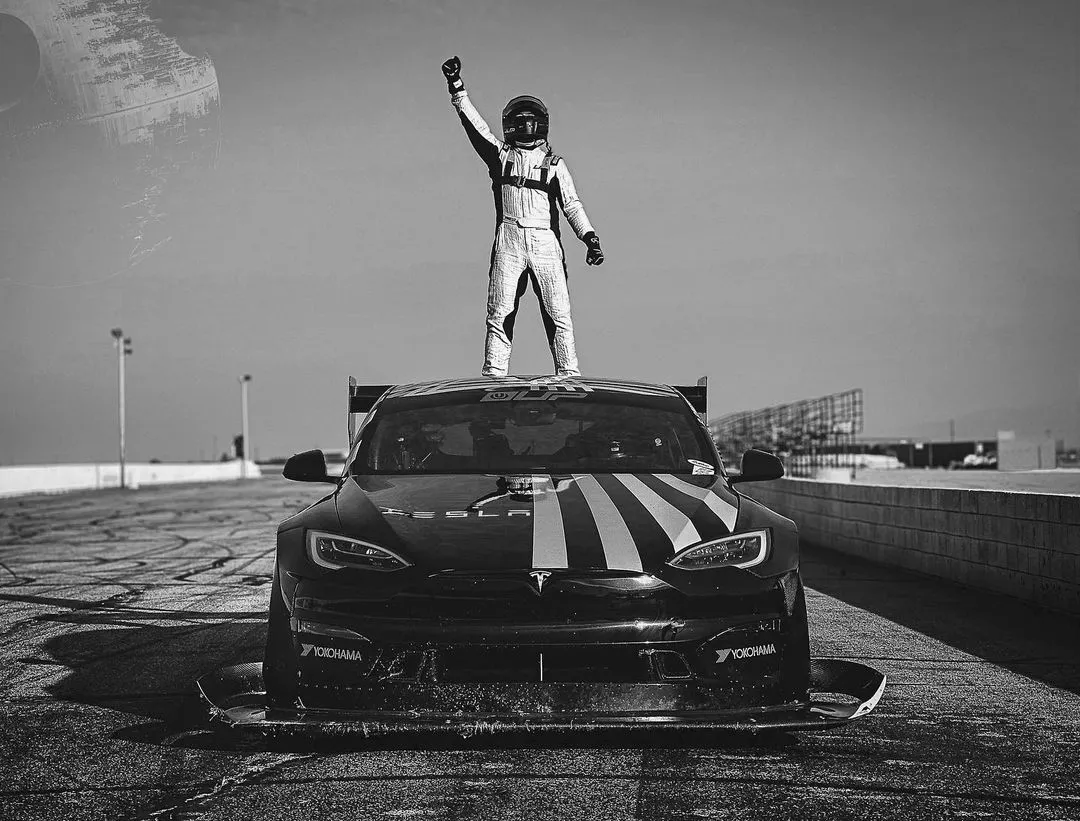 Racing driver in full gear standing triumphantly on the roof of a Tesla race car on a racetrack.